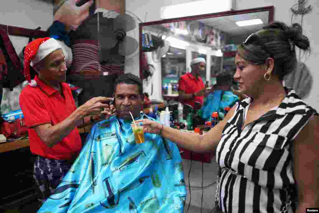 René Sacarras, con gorro navideño, corta el pelo al cuentapropista Yaislan Alonso mientras bebe un trago de la mano de su esposa en el centro de La Habana, el 9 de diciembre de 2023. REUTERS/Alexandre Meneghini