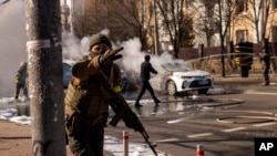 Soldados ucranianos toman posiciones en torno a una instalación militar mientras arden dos automóviles en una calle en Kiev, Ucrania, sábado 26 de febrero de 2022. (AP Foto/Emilio Morenatti)